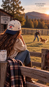 Woman wearing a Foxboro Farms hat beside a pasture fence at sunset, with autumn trees and mountains in the background